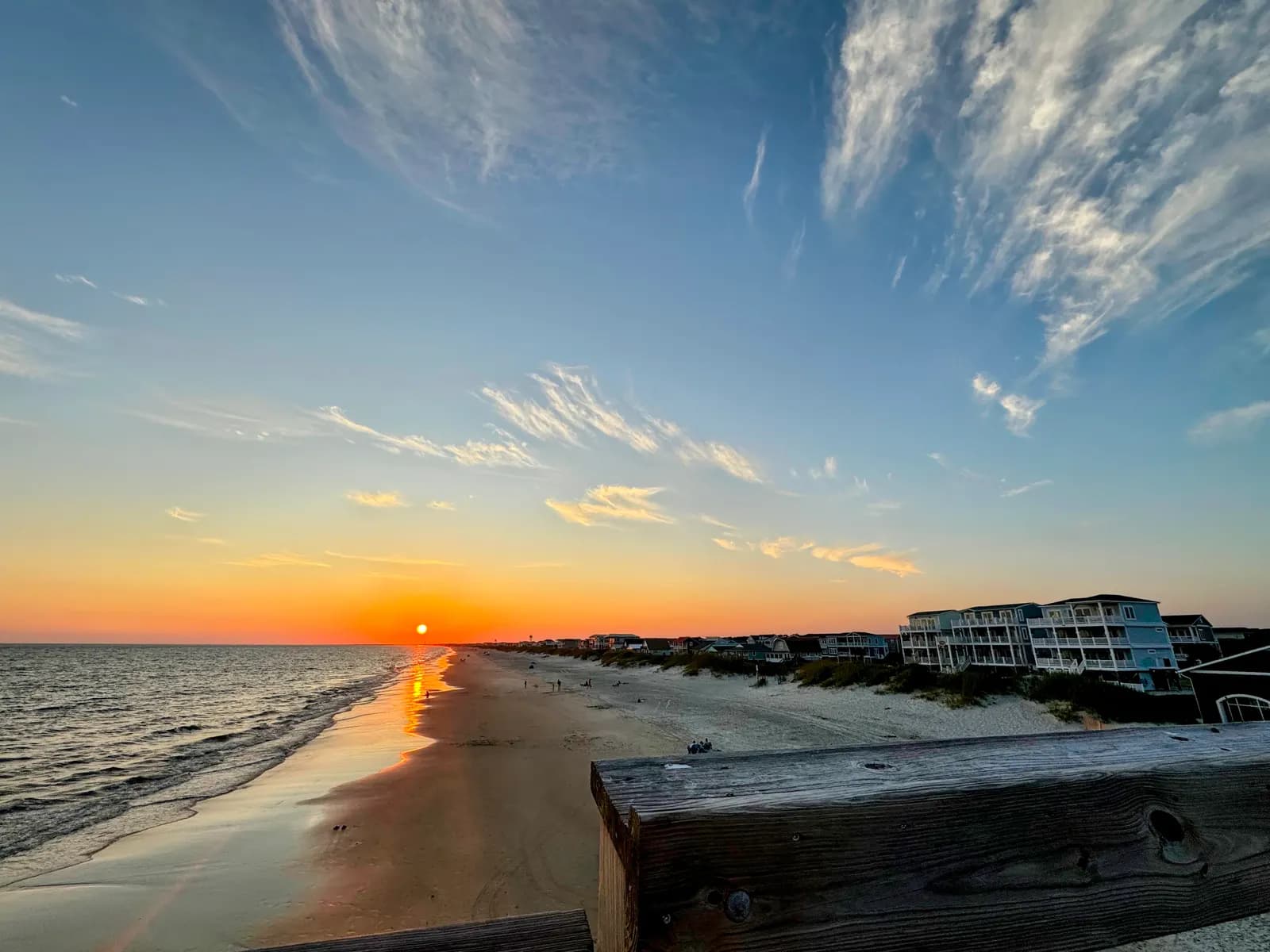 Sunset over the ocean seen from a wooden railing above the beach, with raised beach houses along the dunes.