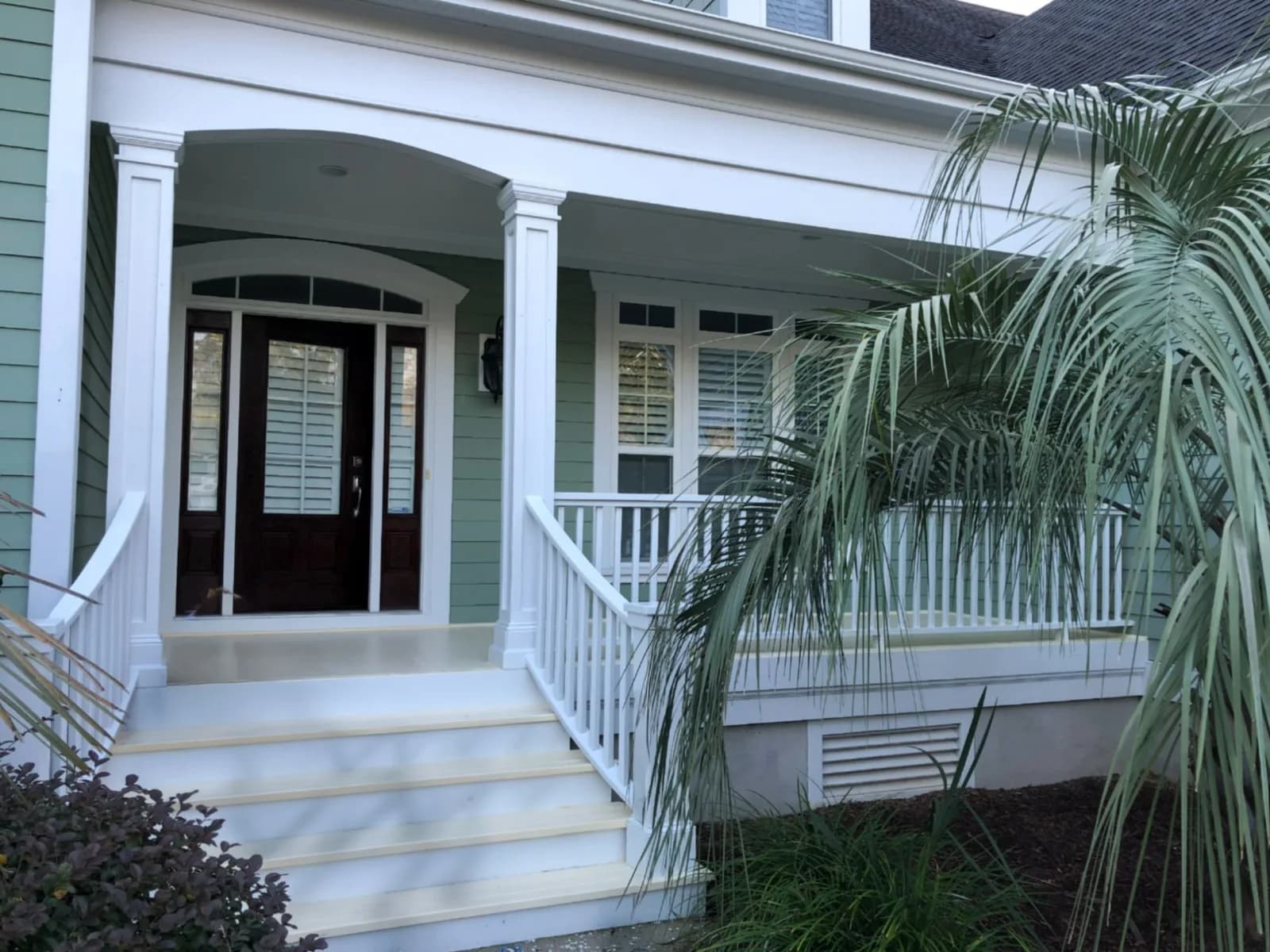 A freshly repainted front porch and steps: a dark front door with sidelights and an arched transom, white square columns, railing and trim against sage-green lap siding, with a palm leaning across the porch.